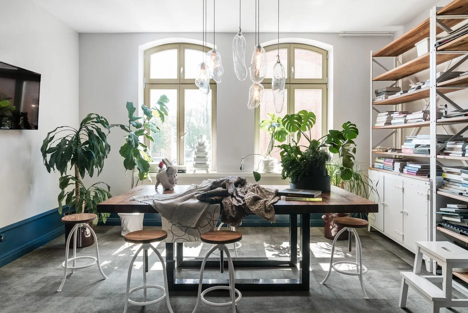 Rustykalny stół jadalny w jasnym pomieszczeniu z roślinami, lampami wiszącymi i przemysłowymi regałami w aranżacji wnętrz w Gdańsku